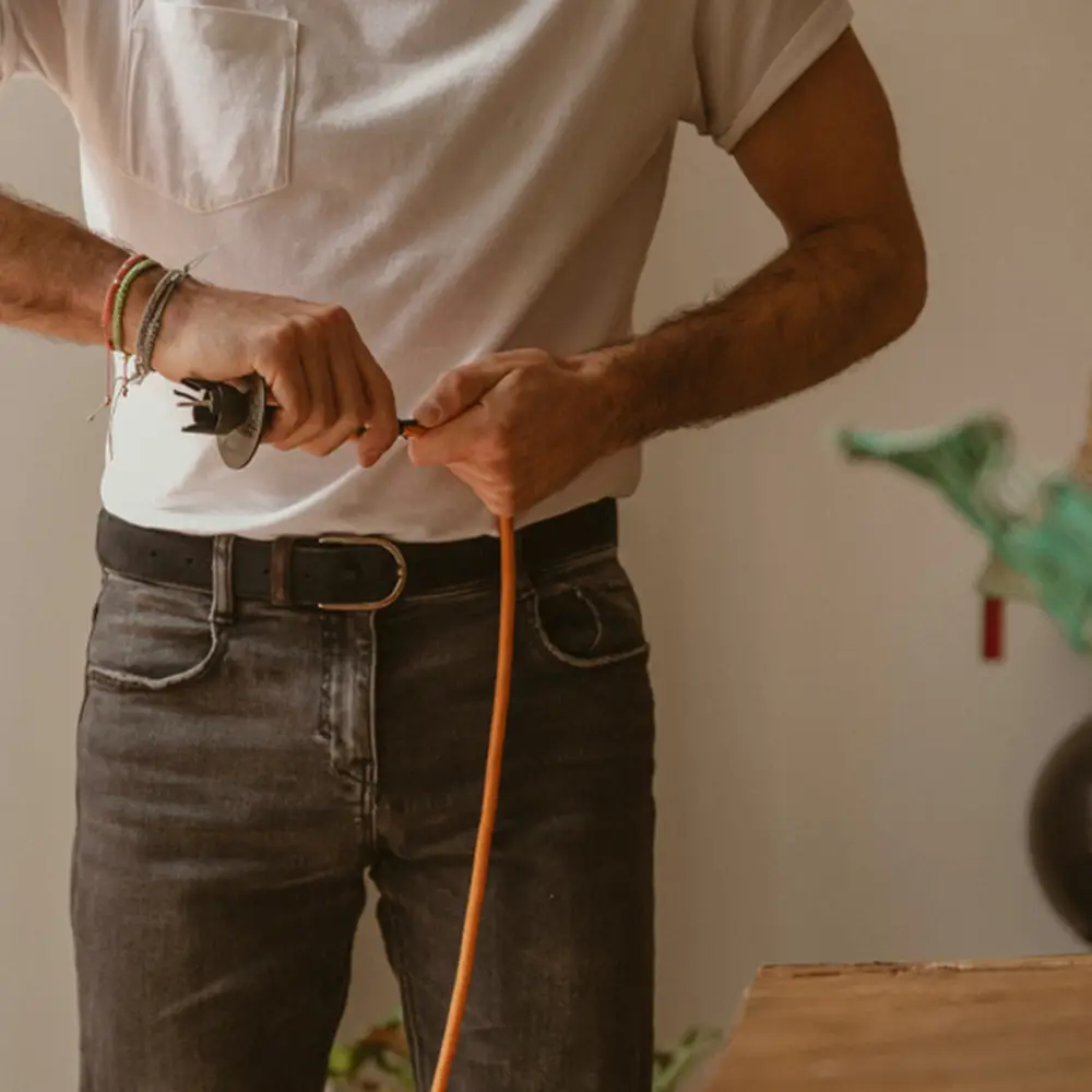 a male person holding an orange wire cable wearing black denim jeans and a white tee standing in front of a beige wall next to a wooden table with a plant on it